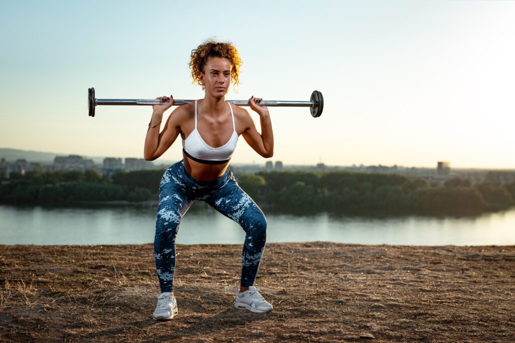 Young fitness woman doing workout with barbell by the river in a sunset.
